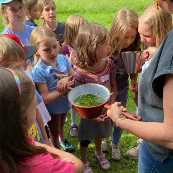 Eine Gruppe von Kindern und einem Erwachsenen sind im Freien, der Erwachsene hält eine Schüssel mit grünen Blättern, während die Kinder aufmerksam zuschauen.