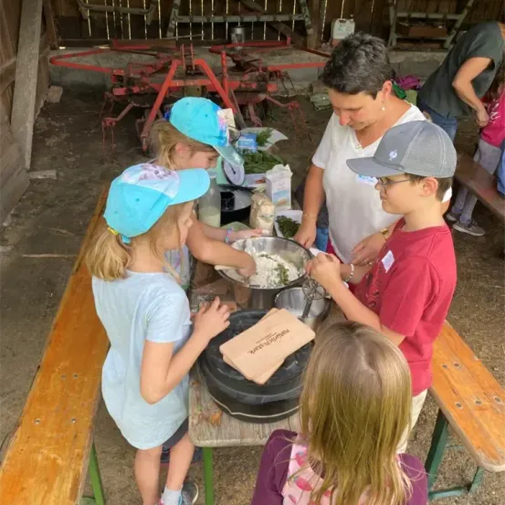 Eine Gruppe von Kindern und einem Erwachsenen sind in einer Scheune und bereiten Essen zu. Die Kinder tragen Mützen und der Erwachsene trägt ein weißes Hemd. Die Kinder stehen um einen Tisch mit Schüsseln und Tüten.