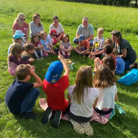 Eine Gruppe von Kindern und Erwachsenen sitzt im Kreis auf einem Grasfeld, einige halten Essen. Ein Kind zeigt, während andere zuschauen. Die Erwachsenen, darunter eine Frau mit Sonnenbrille, führen eine Unterhaltung.