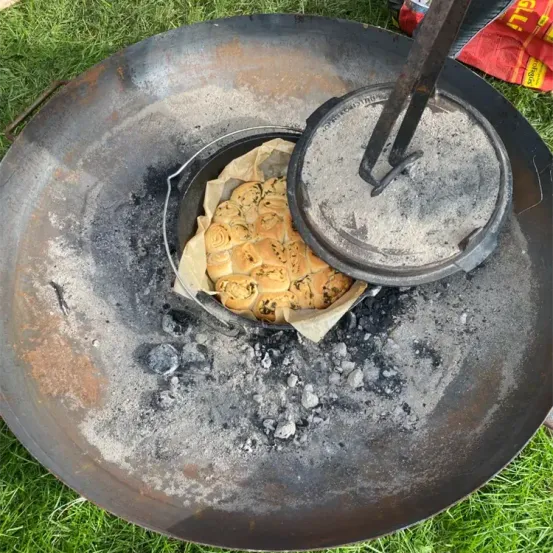 Ein runder Gusseisentopf mit Brötchen steht auf einem Feuerstellen mit Asche auf einer Rasenfläche.