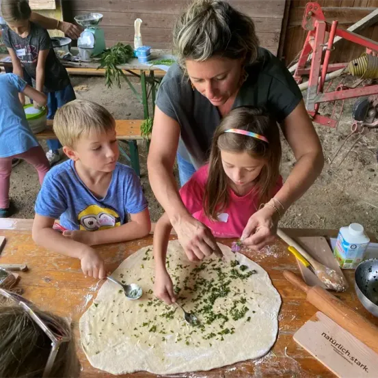 Eine Frau und zwei Kinder bereiten Pizza auf einem Holztisch zu. Die Frau hilft den Kindern, Zutaten auf den Pizzateig zu legen. Eine Schüssel, ein Löffel und ein Nudelholz liegen auf dem Tisch. Dahinter sind weitere Kinder, ein Tisch und eine rote Struktur.