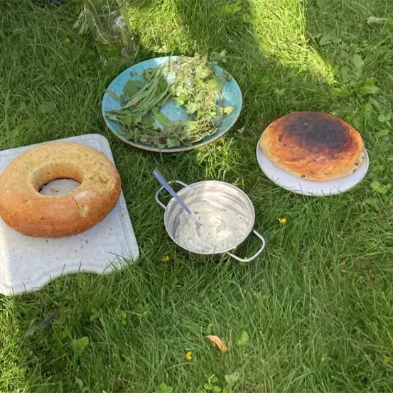An einem sonnigen Tag besteht ein Picknick-Arrangement aus einem runden Brot, einer Schüssel mit Löffel, einem Teller mit verbranntem Gebäck und einem Teller mit grünen Kräutern.