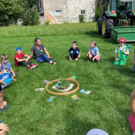 Eine Frau und mehrere Kinder sitzen in einem Kreis auf einem Rasen mit einem Traktor und einem Gebäude im Hintergrund.