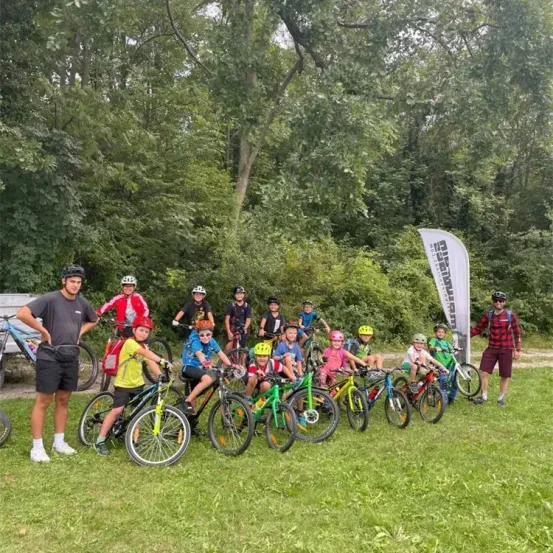 Eine Gruppe von Kindern und zwei Erwachsenen posiert mit ihren Fahrrädern auf einer Rasenfläche mit Bäumen im Hintergrund.