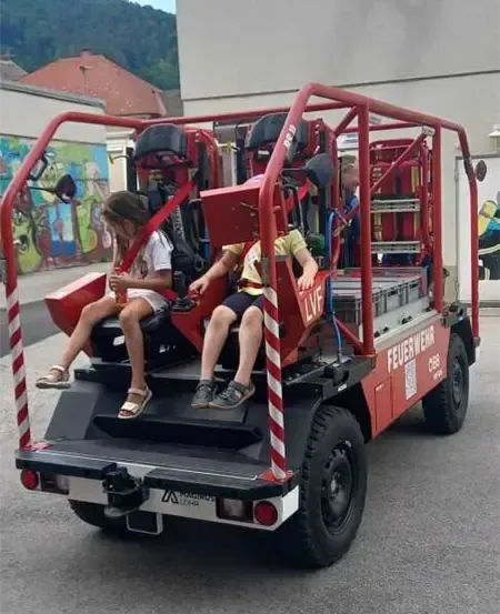 Zwei Kinder fahren einen roten Feuerwehrwagen. Der Wagen steht vor einem Gebäude mit Graffiti. Der Wagen ist mit 'Feuerwehr' und 'LVF' beschriftet.