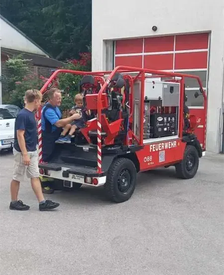 Ein Feuerwehrwagen mit rotem Korpus ist vor einem Gebäude geparkt. Ein Kind sitzt auf dem Fahrersitz. Ein Mann hält die Hand des Kindes.