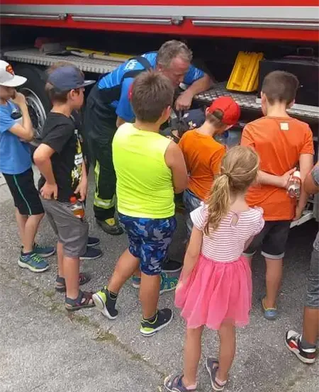 Ein Feuerwehrmann zeigt Kindern, wie man einen Feuerlöscher benutzt. Sie stehen um einen Feuerwehrwagen herum.