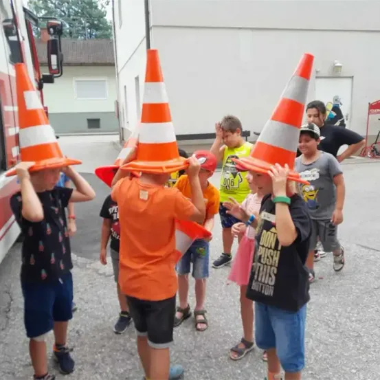 Eine Gruppe von Kindern, die orangefarbene Verkehrskegel tragen und um einen Feuerwehrwagen herumstehen. Dahinter ein Gebäude mit Fenster und Tür.