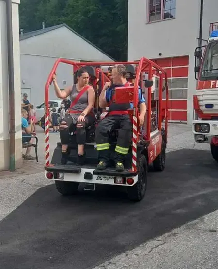 Zwei Personen sitzen auf einem roten Fahrzeug, möglicherweise einem Feuerwehrauto, vor einem Gebäude. Leute stehen am Straßenrand.