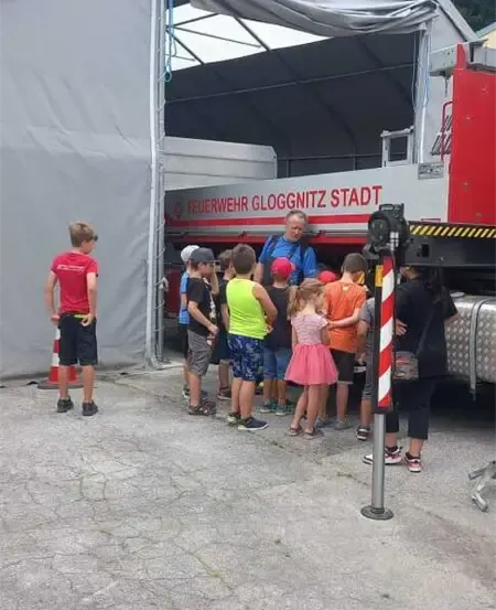 Kinder und Erwachsene stehen vor einem roten Feuerwehrauto mit den Worten 'Feuerwehr Gloggnitz Stadt'. Ein Mann erklärt ihnen etwas.