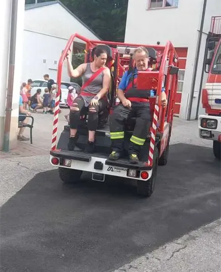 Zwei Personen fahren auf einem roten Feuerwehrwagen vor einem Gebäude mit dem Datum 1863. In der Nähe sitzen mehrere Personen auf einer Bank.