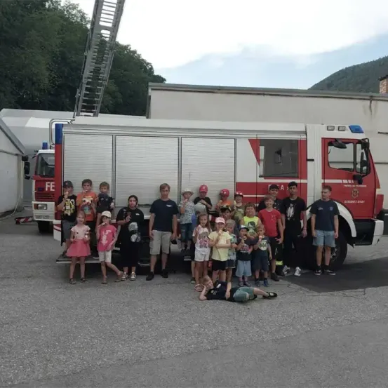 Eine Gruppe von Kindern und Erwachsenen steht vor einem roten Feuerwehrauto mit einer Leiter oben drauf. Sie tragen alle Mützen und einige haben Sonnenbrillen.