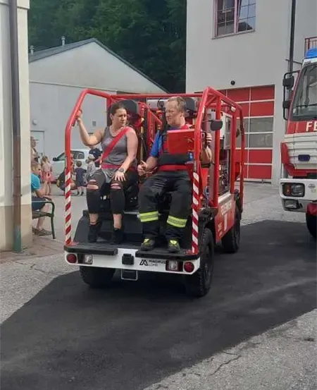 Zwei Personen sitzen auf einem roten Fahrzeug, wahrscheinlich einem Feuerwehrwagen, vor einer Feuerwache.