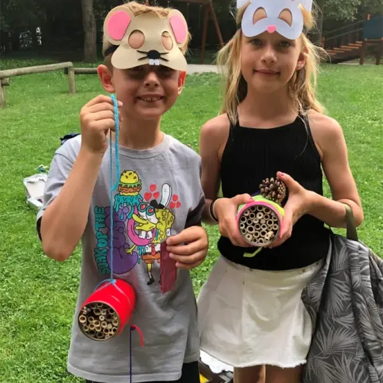 Zwei Kinder, ein Junge und ein Mädchen, sind in einem Park und tragen Masken, während sie Insektenhotels halten. Der Junge hat eine Mausmaske und eine rote Röhre, während das Mädchen eine Katzenmaske und eine bunte Röhre hat.