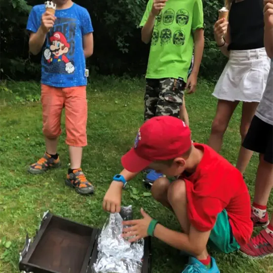 Mehrere Kinder versammeln sich um eine offene Box, möglicherweise beim Kochen. Ein Kind, das einen roten Hut trägt, kniet und interagiert mit dem Inhalt. Andere halten Eiswaffeln. Die Umgebung ist im Freien auf Gras, mit Bäumen im Hintergrund.