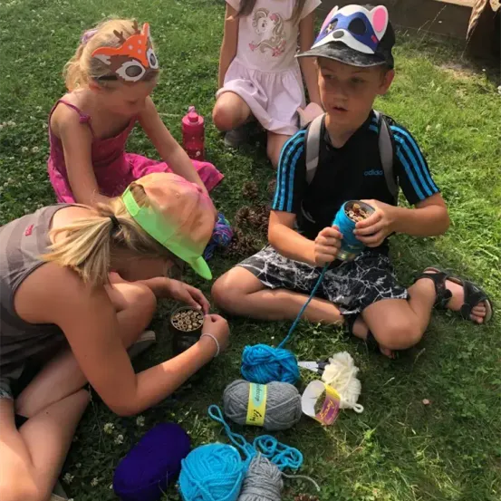 Kinder sitzen auf dem Gras, zwei mit Masken und eines mit einem Hut. Ein Mädchen strickt mit Garn, während ein Junge eine Dose hält.