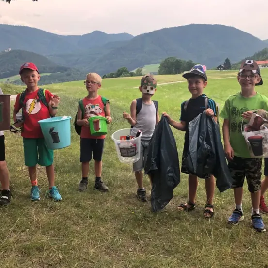 Eine Gruppe von Kindern steht auf Gras, trägt Hüte und Brillen, hält Eimer und trägt Rucksäcke. Einer trägt eine Gesichtsmaske. Sie sind auf einem Feld mit Bergen im Hintergrund.