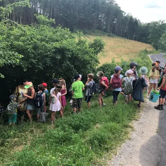 Eine Gruppe von Kindern steht in einer Reihe am Straßenrand, einige halten Taschen und andere tragen Hüte und haben Rucksäcke.