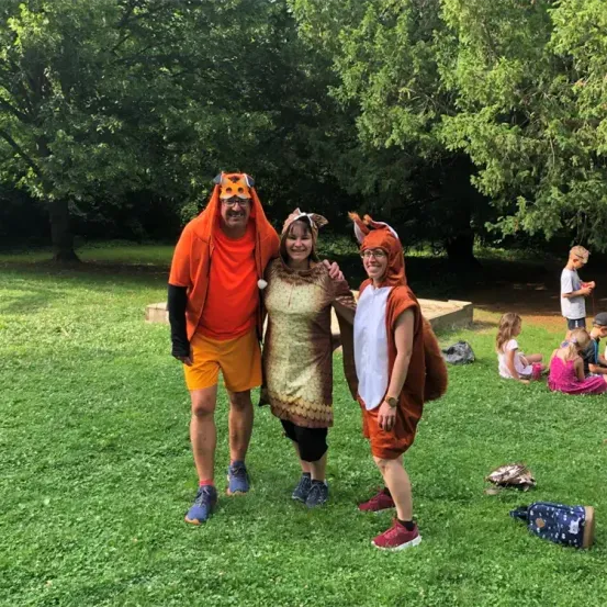 Drei Menschen in Tierkostümen stehen in einem Park. Ein Junge in einem weißen Hemd geht neben ihnen. Mehrere Kinder sitzen hinter ihnen auf dem Gras.