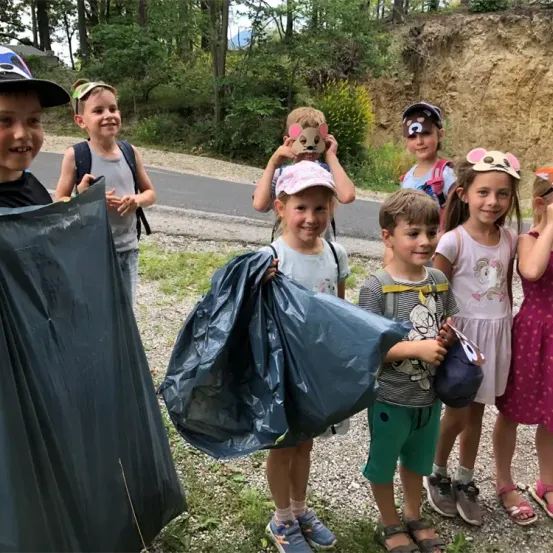 Eine Gruppe von Kindern mit Tiermasken steht auf Kies und hält Müllsäcke in der Hand, lächelt. Ein Kind hält einen blauen Plastiksack. Sie sind in der Nähe eines Erdhügels mit Bäumen.