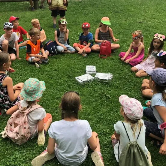 Eine Gruppe von Kindern sitzt im Kreis auf einem Grasfeld, einige mit Rucksäcken und Mützen. Ein Kind hält eine rote Tasche, und in der Mitte befinden sich Folienverpackungen.