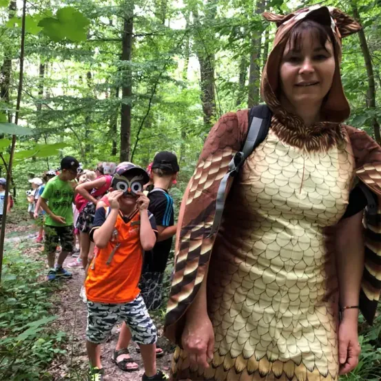Eine Gruppe von Kindern und eine erwachsene Frau in einem Kostüm wandern im Wald. Die Frau trägt ein Eulenkostüm und die Kinder haben Mützen und Turnschuhe an.