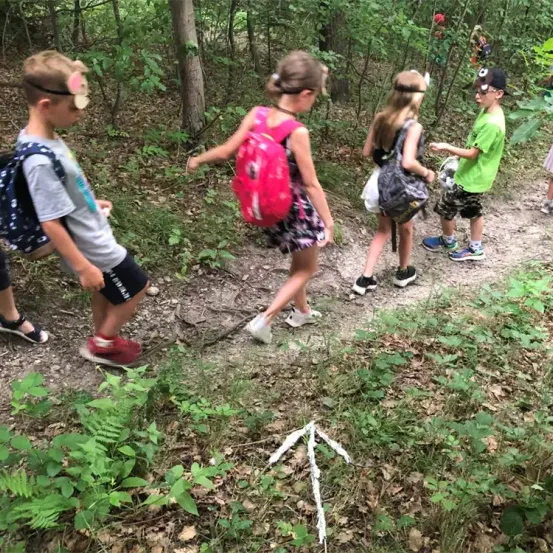 Vier Kinder, die Rucksäcke tragen, wandern auf einem Waldpfad. Sie tragen Schuhe und haben verschiedene Accessoires wie Hüte und Brillen. Das Mädchen in der Mitte trägt einen rosa Rucksack.