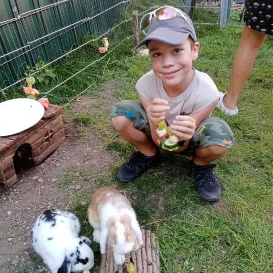 Ein Junge mit Hut und Turnschuhen hockt auf Gras und füttert zwei Kaninchen mit Gemüse. Dahinter ist eine Holzstruktur mit einem Teller und einem Zaun zu sehen.