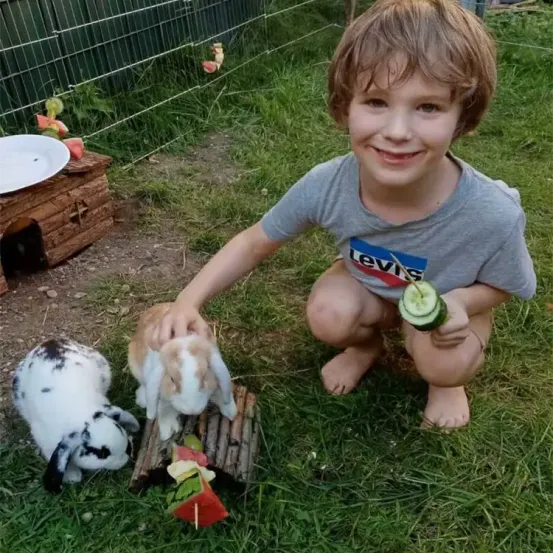 Ein junger Junge lächelt, während er auf dem Gras kniet und zwei Kaninchen mit einer Gurke und Wassermelone füttert. Ein hölzernes Kaninchenhaus und ein Zaun sind im Hintergrund.
