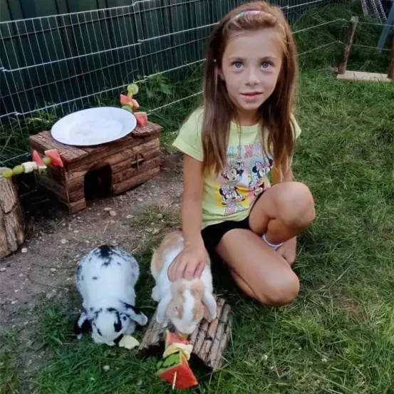 Ein junges Mädchen sitzt auf dem Gras und füttert zwei Kaninchen mit Wassermelonen-Spießen. Hinter ihr steht ein Holzhütte mit einem Teller oben und einem eingezäunten Bereich.