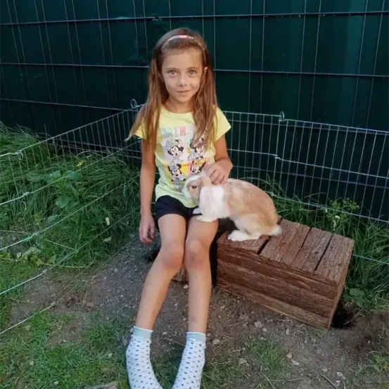 Ein Mädchen mit langen Haaren sitzt auf einem Holzhocker in einem Grasbereich. Sie hält einen Hasen. Hinter ihr befindet sich ein Käfig mit einem grünen Metallzaun.