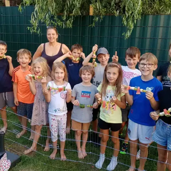Eine Gruppe von Kindern und eine erwachsene Frau posieren für ein Foto. Sie alle haben Wassermelonenstäbchen in den Händen. Sie stehen in einem Garten mit einem grünen Zaun.
