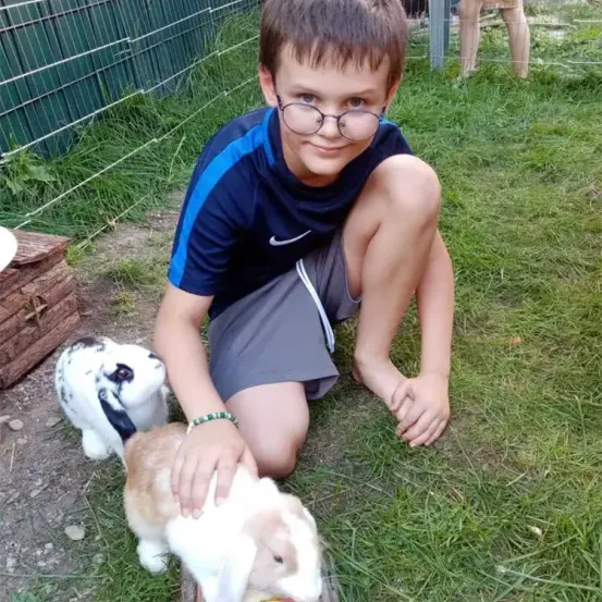 Ein Junge mit Brille hält ein kleines Kaninchen, während ein anderes Kaninchen zusieht. Sie befinden sich in einem eingezäunten Außenbereich.