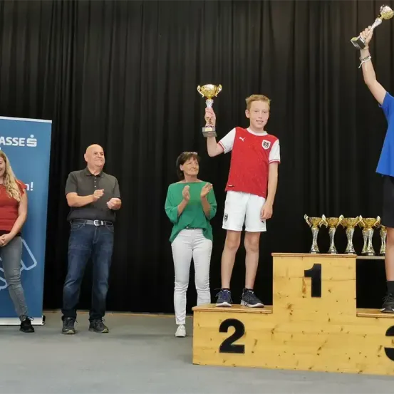 Ein junger Junge steht auf einem Podest und hält einen Pokal in der Hand, flankiert von zwei Erwachsenen. Eine weitere Person hält im Hintergrund einen Pokal. Ein Banner mit weißem Text ist sichtbar.