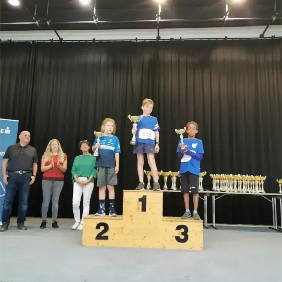 Drei Kinder stehen auf einem Podest und halten Trophäen. Hinter ihnen applaudieren mehrere Erwachsene. Auf der Bühne stehen zwei Tische mit Trophäen darauf. Hinter der Bühne befindet sich ein schwarzer Vorhang.