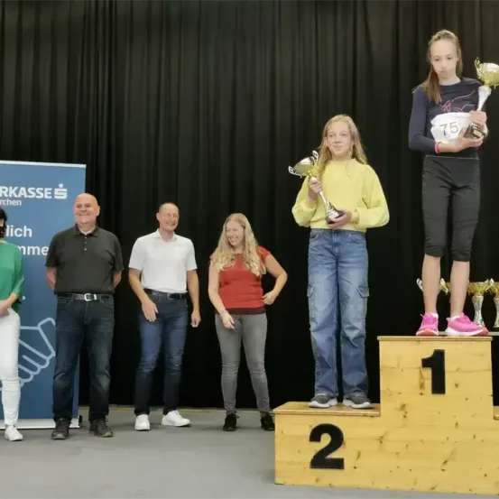 Eine Gruppe von Menschen steht auf einer Bühne, wahrscheinlich bei einer Preisverleihung. Eine Frau steht auf dem Siegertreppchen und hält eine Trophäe, während eine andere Frau auf dem zweiten Platz steht und eine Trophäe hält. Andere Personen stehen hinter ihnen, und ein Banner mit Text ist sichtbar.