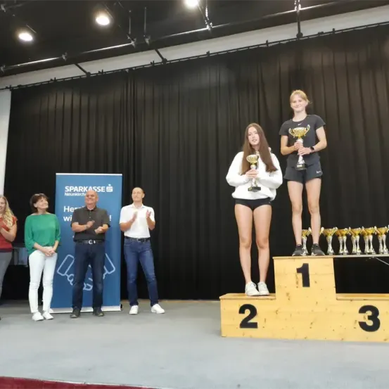 Zwei Mädchen stehen mit Pokalen auf einem Podest, begleitet von zwei Männern und zwei Frauen. Ein blauer Banner mit Text ist im Hintergrund sichtbar.