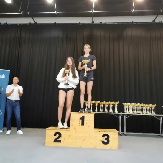 Zwei Frauen stehen auf einem Podium mit Trophäen in der Hand. Sie sind von zusätzlichen Trophäen umgeben. Ein Mann klatscht hinter ihnen. Der Hintergrund ist schwarz.