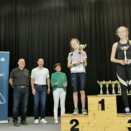 Zwei junge Frauen stehen auf einem Podest und halten Trophäen, während drei Erwachsene hinter ihnen stehen. Einer der Erwachsenen trägt ein grünes Shirt, und zwei tragen weiße Shirts. Sie alle lächeln.