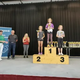 Drei Mädchen stehen auf einem Podest und halten Trophäen. Das Mädchen in der Mitte hat einen Nummer-32-Anhänger an ihrem Trikot. Hinter ihnen stehen einige Leute, und ein Tisch ist mit Trophäen gefüllt. Ein blauer Banner mit Text steht auf der linken Seite.