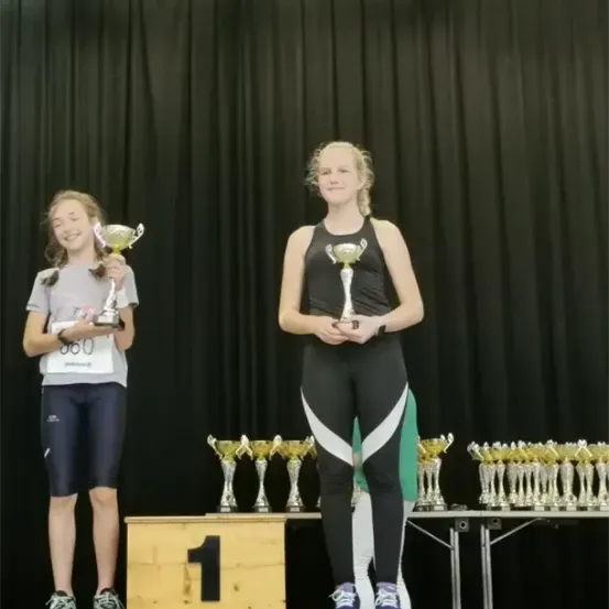 Zwei Mädchen stehen mit Trophäen auf einem Podest und lächeln. Dahinter sind mehrere Trophäen auf einem Tisch ausgestellt. Das Podest ist nummeriert eins, zwei und drei.