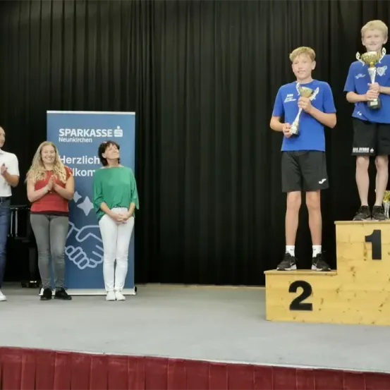 Zwei Jungen stehen auf einer Bühne und halten Pokale in den Händen, während zwei Frauen hinter ihnen klatschen. Ein Junge trägt ein blaues Shirt, der andere ein gelbes. Ein Banner mit dem Wort 'Sparkasse' ist hinter ihnen.