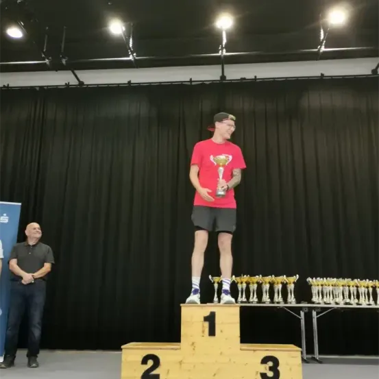 Ein Mann steht auf einem Podest, hält einen Pokal und hat die Zahlen 1, 2 und 3 unter sich. Dahinter sind Trophäen aufgereiht. Ein Mann steht im Hintergrund und ein Banner ist links.