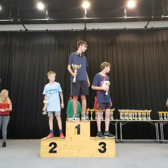 Drei Jungen stehen auf einem Podest mit Trophäen. Der Junge in der Mitte trägt Kopfhörer. Hinter ihnen stehen viele Trophäen. Eine Frau steht links.