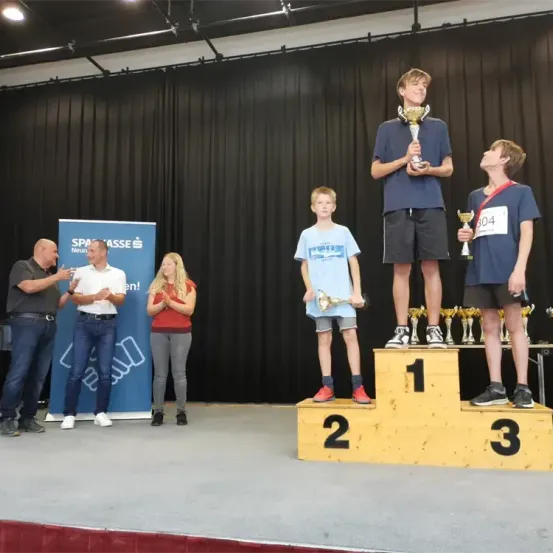 Drei Jungen stehen auf einem Podest mit den Nummern 1, 2 und 3. Der Junge auf der linken Seite hält einen Pokal und trägt eine rote Medaille. Der Junge in der Mitte hält einen Pokal. Der Junge auf der rechten Seite steht und hält einen Pokal. Hinter ihnen klatschen zwei Männer und eine Frau. Ein blauer Banner mit dem Text 'SPASSSE' ist hinter ihnen.