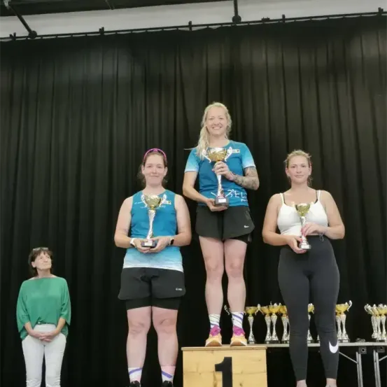 Drei Frauen stehen auf einer Bühne und halten Pokale. Die Frau in der Mitte trägt ein blaues Shirt und hat eine Tätowierung am Arm. Die anderen beiden Frauen tragen schwarze Leggings und halten Pokale. Hinter ihnen steht ein schwarzer Vorhang mit Pokalen.