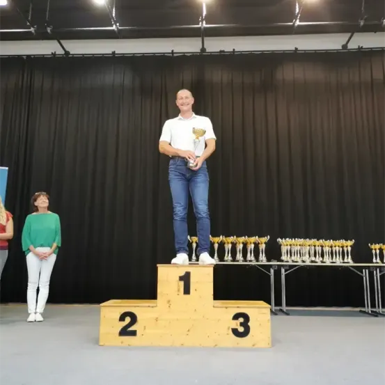Ein Mann steht auf der obersten Stufe eines Podests, hält eine Trophäe und lächelt, während mehrere Trophäen auf einem Tisch dahinter angeordnet sind. Eine Frau steht in der Nähe und schaut ihn an.