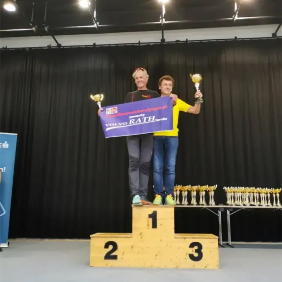 Zwei Männer stehen auf einem Podest mit einem blauen Banner, auf dem 'Volvo Rath' steht. Sie halten Pokale und lächeln. Ein Tisch mit weiteren Pokalen steht hinter ihnen. Die Bühne ist mit schwarzen Vorhängen bedeckt.