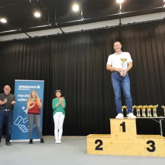 Ein Mann steht lächelnd auf einem Podest und hält eine Trophäe. Dahinter klatschen zwei Frauen. Ein Banner auf der linken Seite sagt 'Sparkasse'. Rechts stehen Trophäen auf einem Tisch.