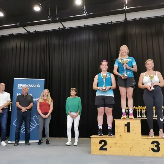 Drei Frauen stehen auf einem Podium, lächeln und halten Trophäen. Zwei Frauen stehen auf der obersten Stufe, eine auf der zweiten, und vier Erwachsene stehen darunter, darunter zwei Männer. Der Hintergrund zeigt ein blaues Banner mit weißem Text.